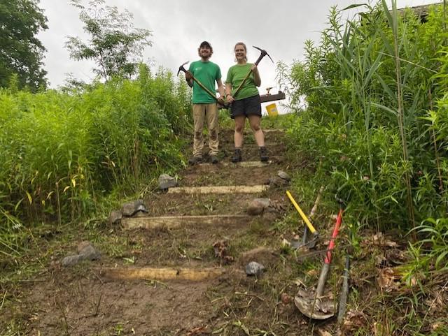 Stella and Jacob stand with tools in their hands amidst a stairwell they've just built.